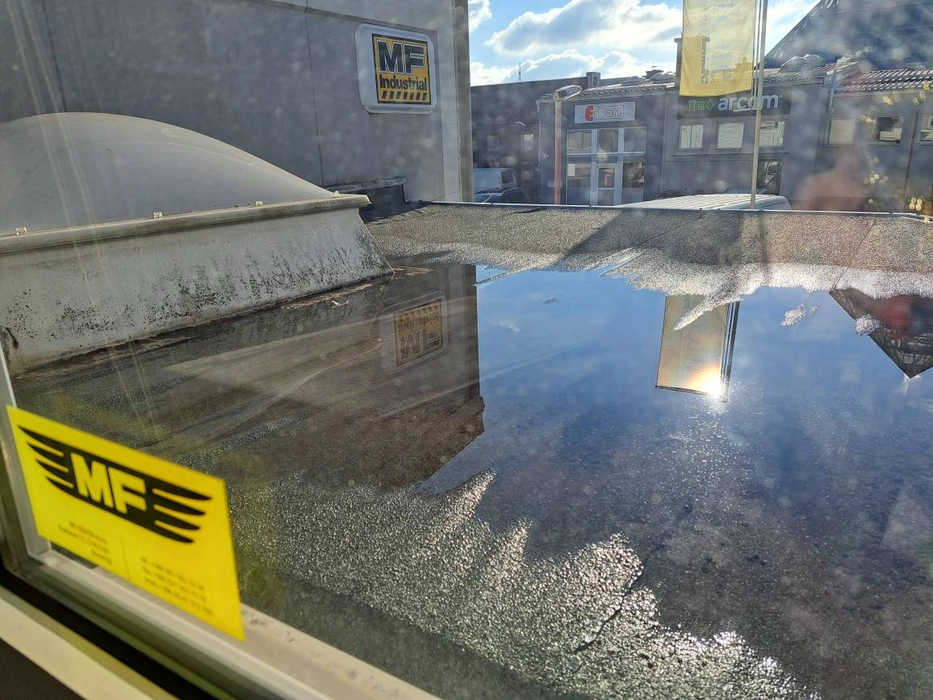 Wasseransammlung auf einem flachen Gewerbedach unter Sonnenlicht, das nahegelegene Gebäude und den Himmel reflektiert.