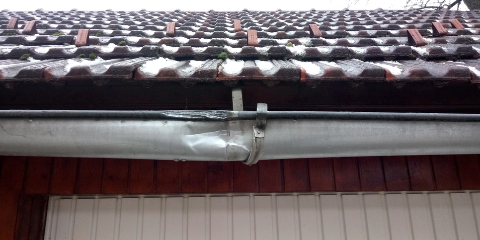 Eine Metallregenrinne ist sichtbar eingedellt unter einem Ziegeldach, was auf mögliche Probleme bei der Wasserableitung hinweist.