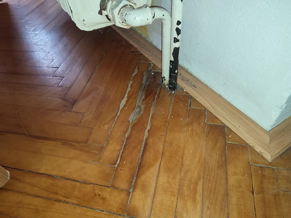 Nahaufnahme eines Holzbodens mit Wasserschaden in der Nähe eines Heizungsrohrs, der verzogene und verfärbte Dielen zeigt.