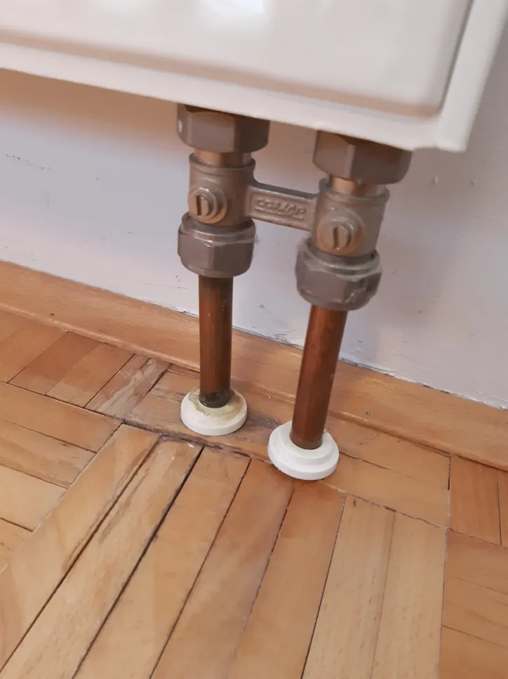 Wasserschaden sichtbar auf dem Holzboden unter den Heizkörperrohren, was auf mögliche Leckagen oder Verschüttungen hinweist.