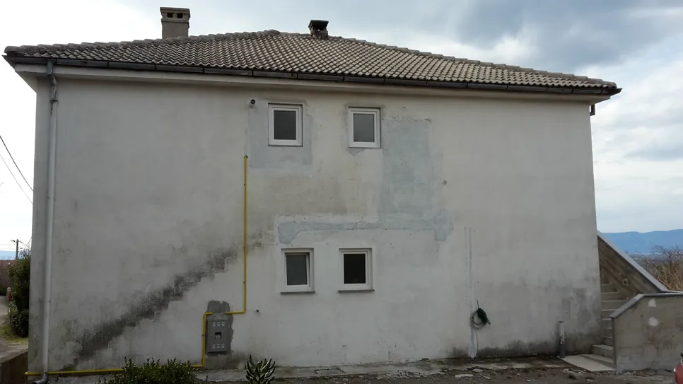 Schimmelwachstum an der außenwand eines hauses Schimmelwachstum sichtbar an der Außenwand eines Hauses unter zwei kleinen Fenstern, was auf Feuchtigkeitsprobleme hinweist.